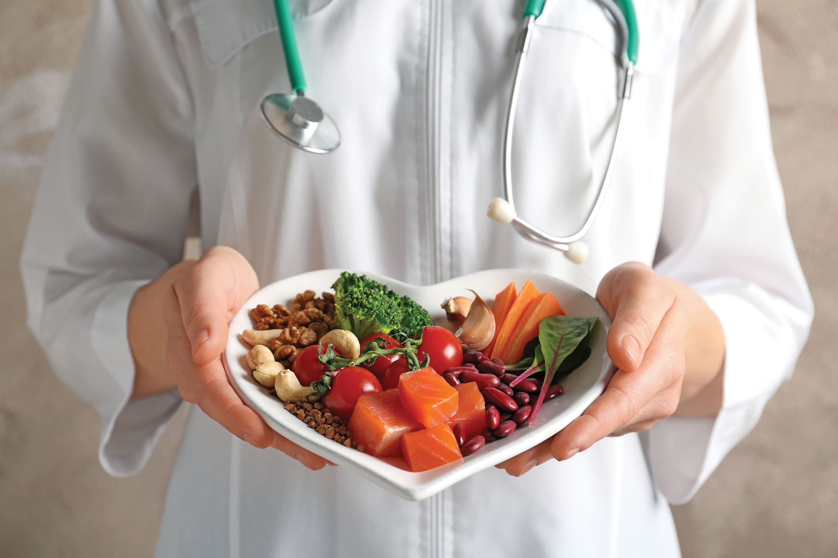 A healthcare professional in a white coat holding a heart-shaped bowl filled with fresh vegetables.