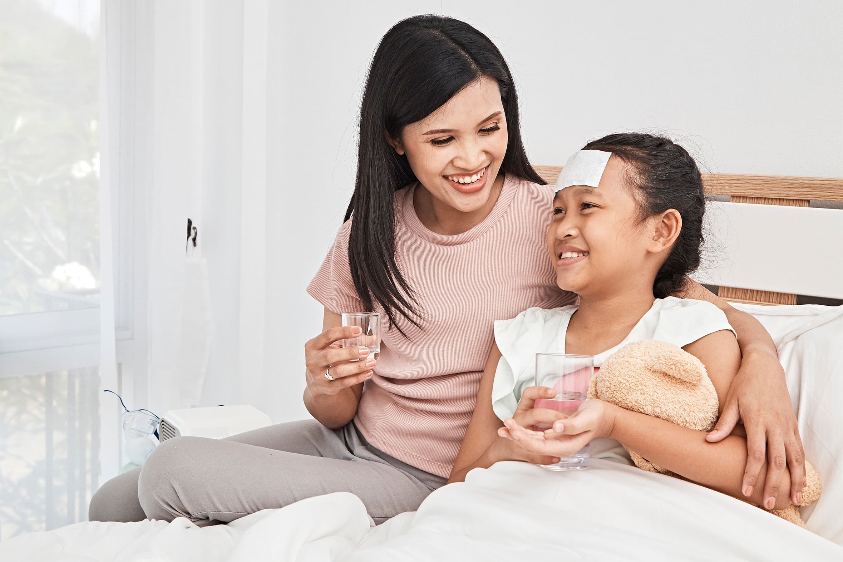 A smiling woman sits on a bed beside a young girl who has a cooling patch on her forehead while holding a glass of water.