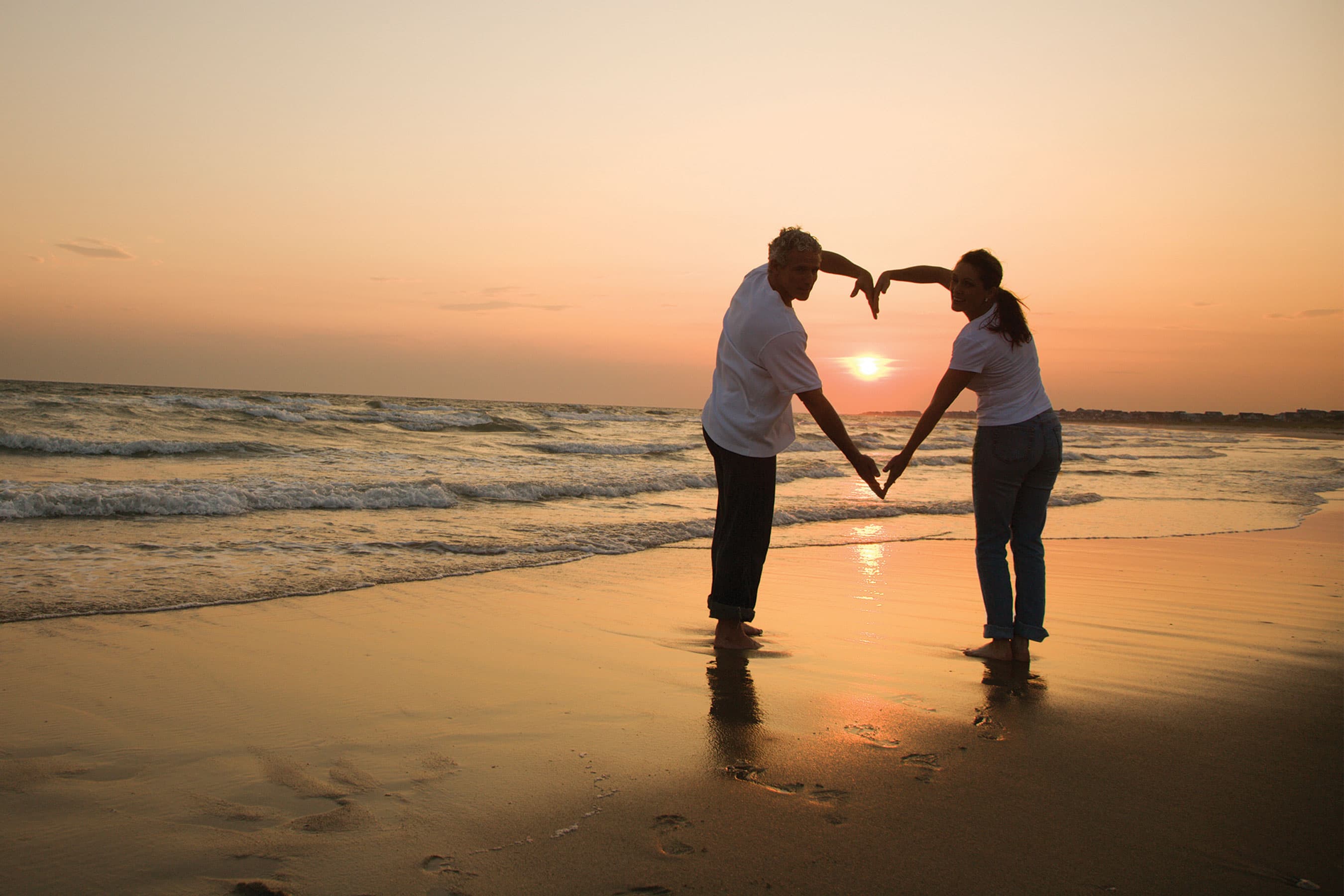 A couple standing on a beach at sunset forming a heart shape with their hands against the setting sun.