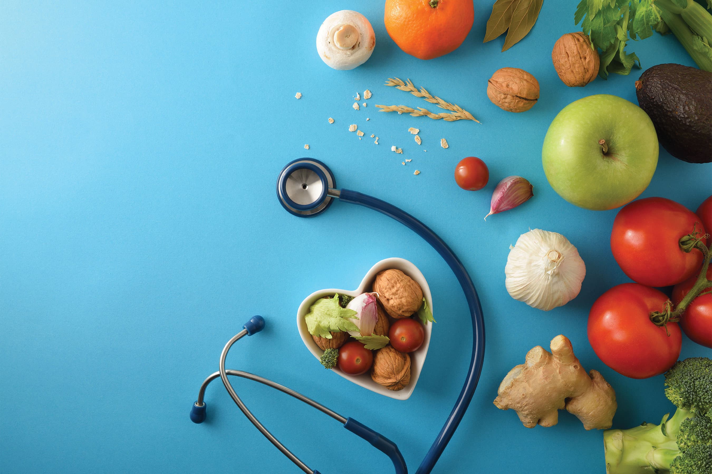 Top-down view of fresh fruits, vegetables, and a heart-shaped bowl surrounded by a stethoscope on a blue surface.