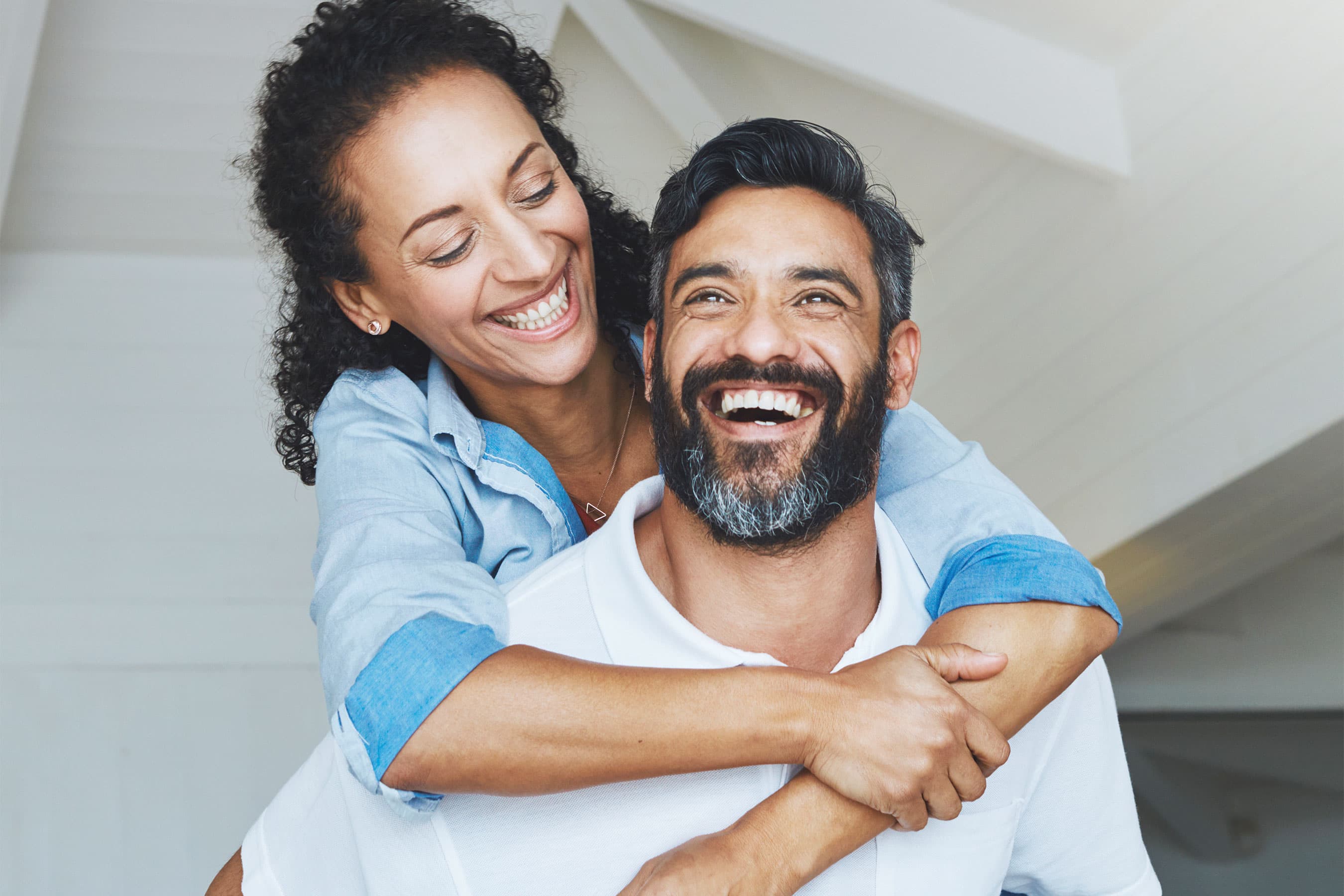 A woman hugging a man from behind as both smile happily indoors.