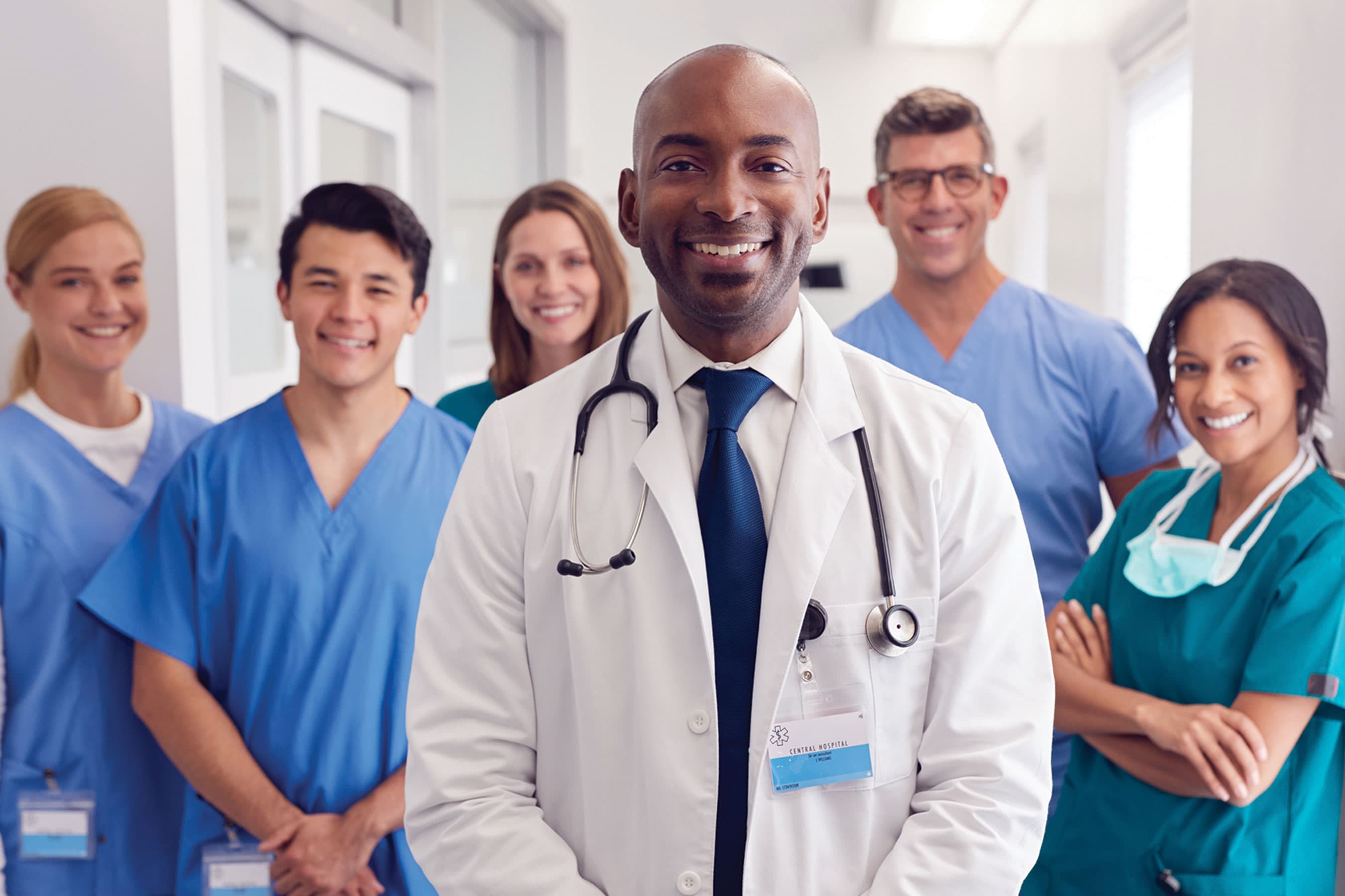 A group of smiling medical professionals and doctors in scrubs and white coats standing in a hallway.